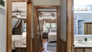 A bedroom with a bed, bedside table, lamp, and a view of a bathroom with a sink and mirror. - Charming Cabin with Fire Pit (Arden)