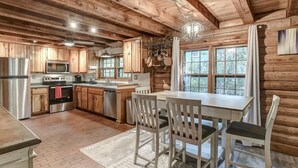 A rustic kitchen with wooden cabinets, a stainless steel refrigerator, a microwave, an oven, and a sink. There is a dining table with chairs and a chandelier hanging above. - Charming Cabin with Fire Pit (Arden)