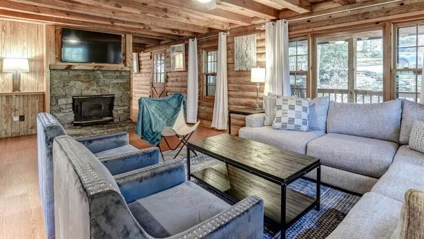 A log cabin interior with a stone fireplace, a ceiling fan, and a large window overlooking a snowy landscape. - Charming Cabin with Fire Pit (Arden)