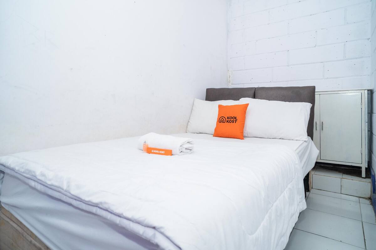 Ein ordentlich bezogenes Bett mit weißer Bettwäsche und einem orangefarbenen Kopfkissen, eine weiße Wand und ein Metallschrank.