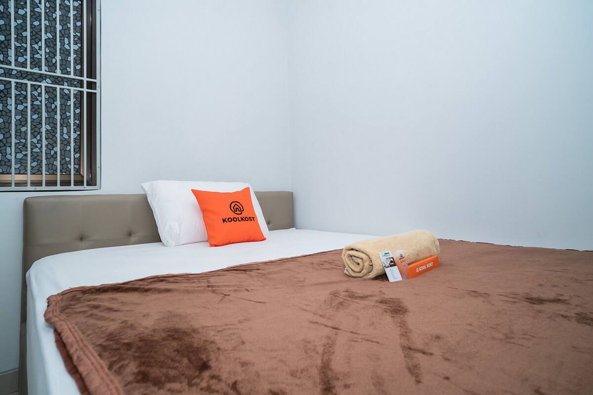 A hotel room with a bed, a white pillow with an orange logo, a brown blanket, and a window with a metal grill.
