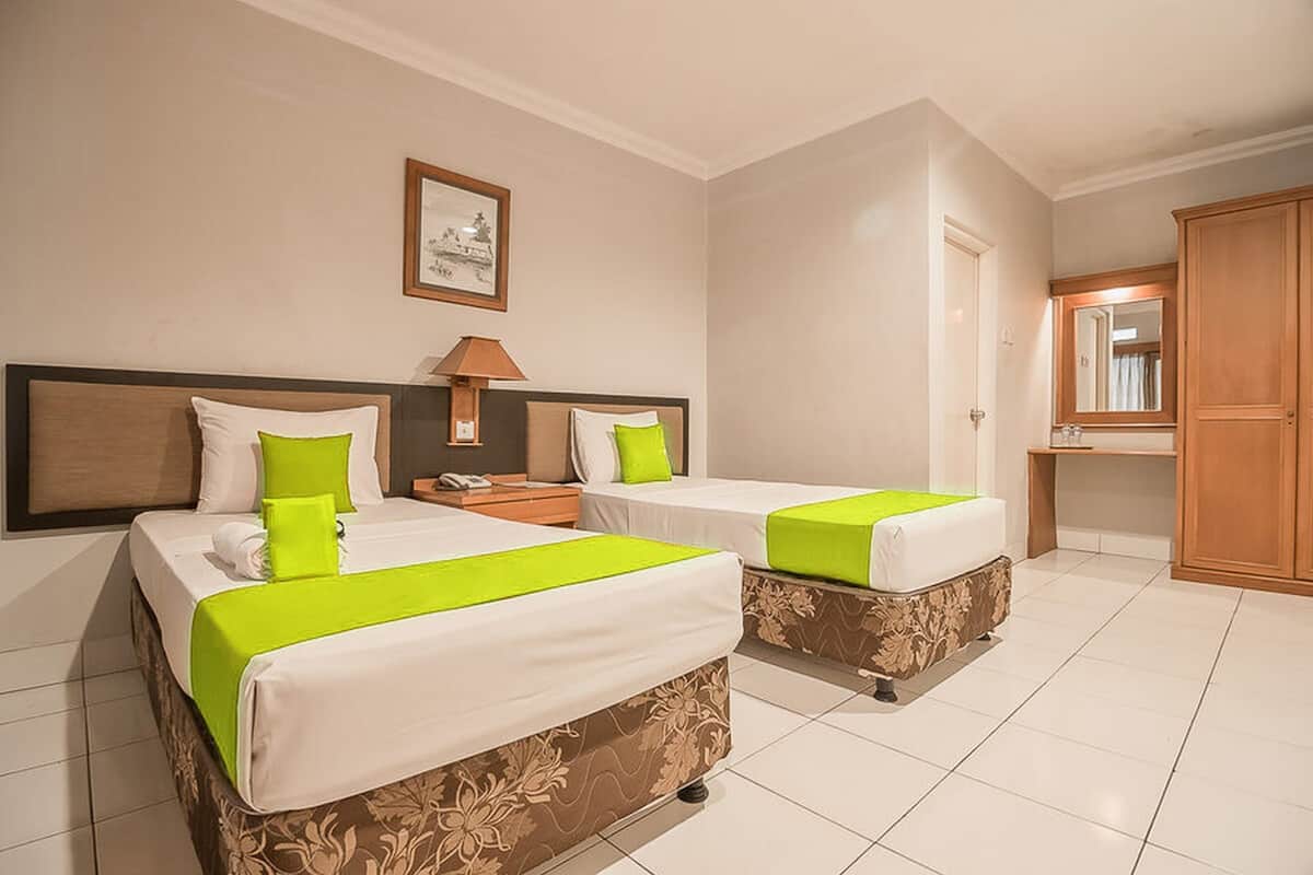 A hotel room with two beds, each with a green bedspread and white pillows, a nightstand with a lamp, a framed picture on the wall, and a wooden wardrobe.