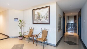 A modern hotel lobby with a polished floor, a framed cityscape artwork, wicker chairs, a potted plant, and a staircase leading to another area. - New Gentala Hotel RedPartner (Medan)