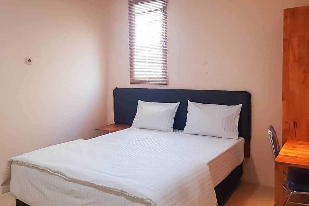 A bedroom with a bed, a nightstand, a wooden desk, and a window with blinds.