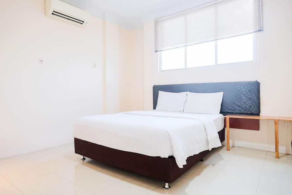 A bedroom with a bed, a bedside table, a window with blinds, and an air conditioning unit.
