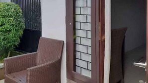 Uma cadeira de vime em frente a uma porta com painel de vidro, um vaso com planta e uma placa com o número da porta.