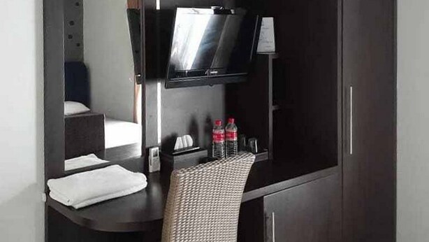 Quarto de hotel compacto com cama, uma mesinha com cadeira, televisão de tela plana na parede e minibar.
