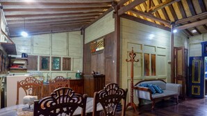 A rustic dining area with wooden chairs and a table, a wooden bench with cushions, and a kitchen area with cabinets and a sink. - Upala Java Huise (Bangunjiwo)