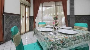 A dining room with a table set for four, chairs with green cushions, a vase with flowers, and a window with curtains. - Hotel Lestari Near Lippo Plaza Mall Jember (Patrang)