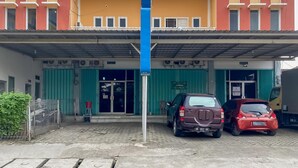 A two-story building with a yellow facade and a blue entrance canopy. There are two cars parked in front, and a green fence to the left. - RedDoorz @ Jalan Morotai Lampung (Bandar Lampung)
