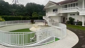 A large, two-story house with a white exterior, a balcony on the upper floor, and a circular garden with a white fence.