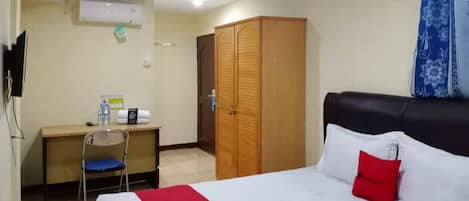 A hotel room with a bed, a desk, a chair, a wardrobe, and an air conditioning unit.