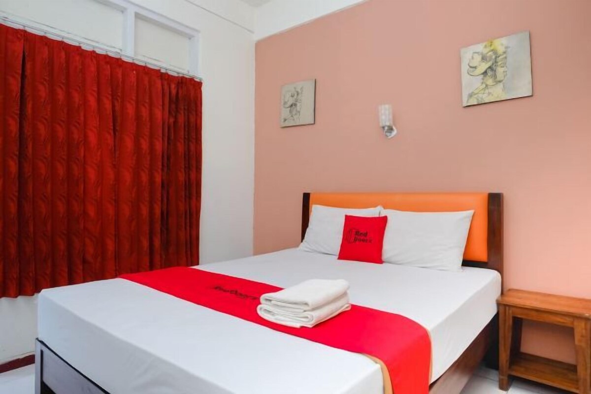 A hotel room with a bed, a wooden nightstand, and a red curtain.