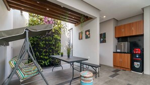 A modern kitchen with wooden cabinets, a stainless steel refrigerator, and a dining area with a swing chair and bench. - RedDoorz Plus near Radio Dalam 3 (Jakarta)