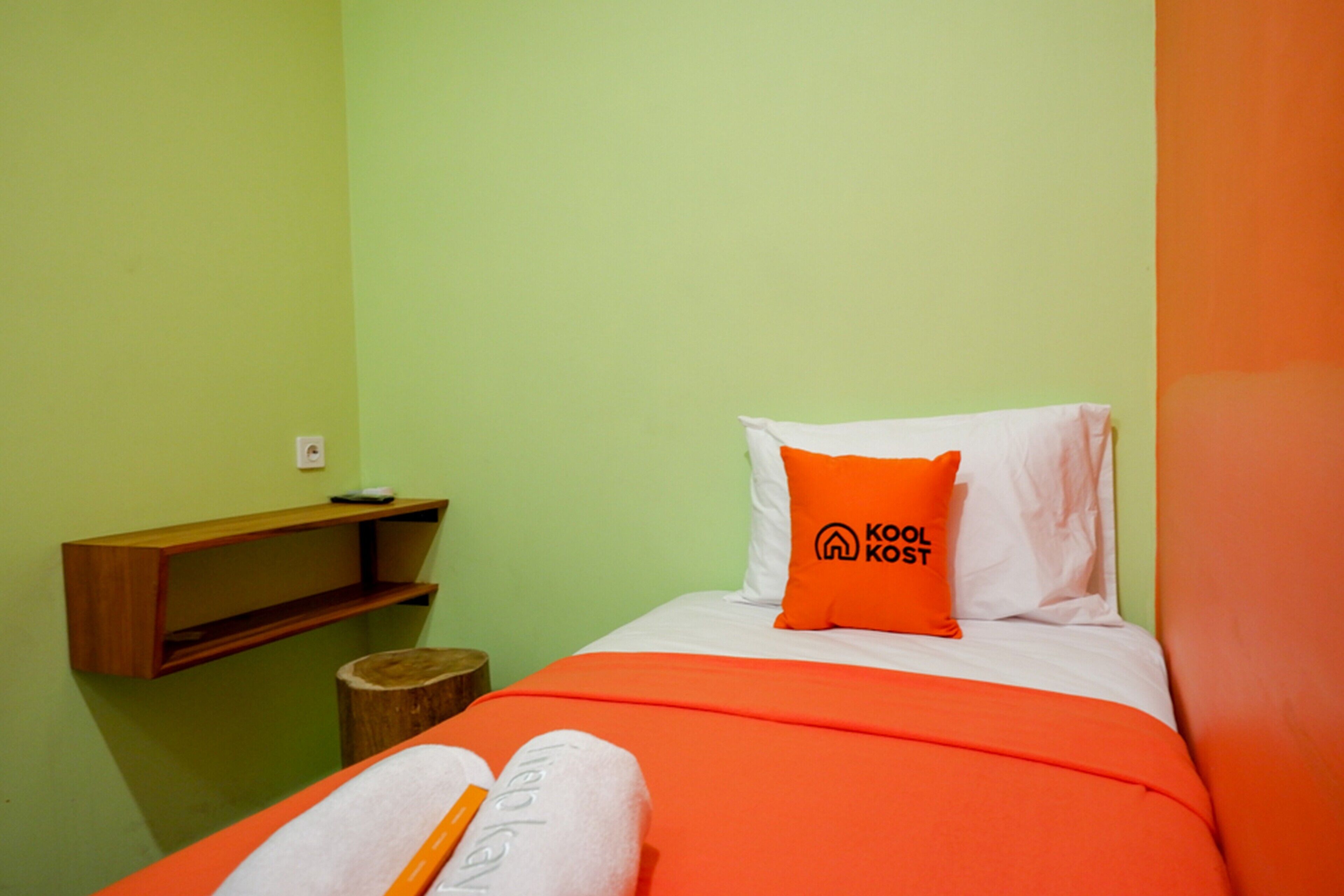 A small, single-bed room with a green wall, a wooden shelf, and a wooden stool.