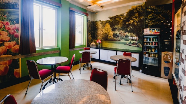 Un restaurante con mesas redondas y sillas rojas, amplios ventanales con cortinas, y un mural que representa un parque.