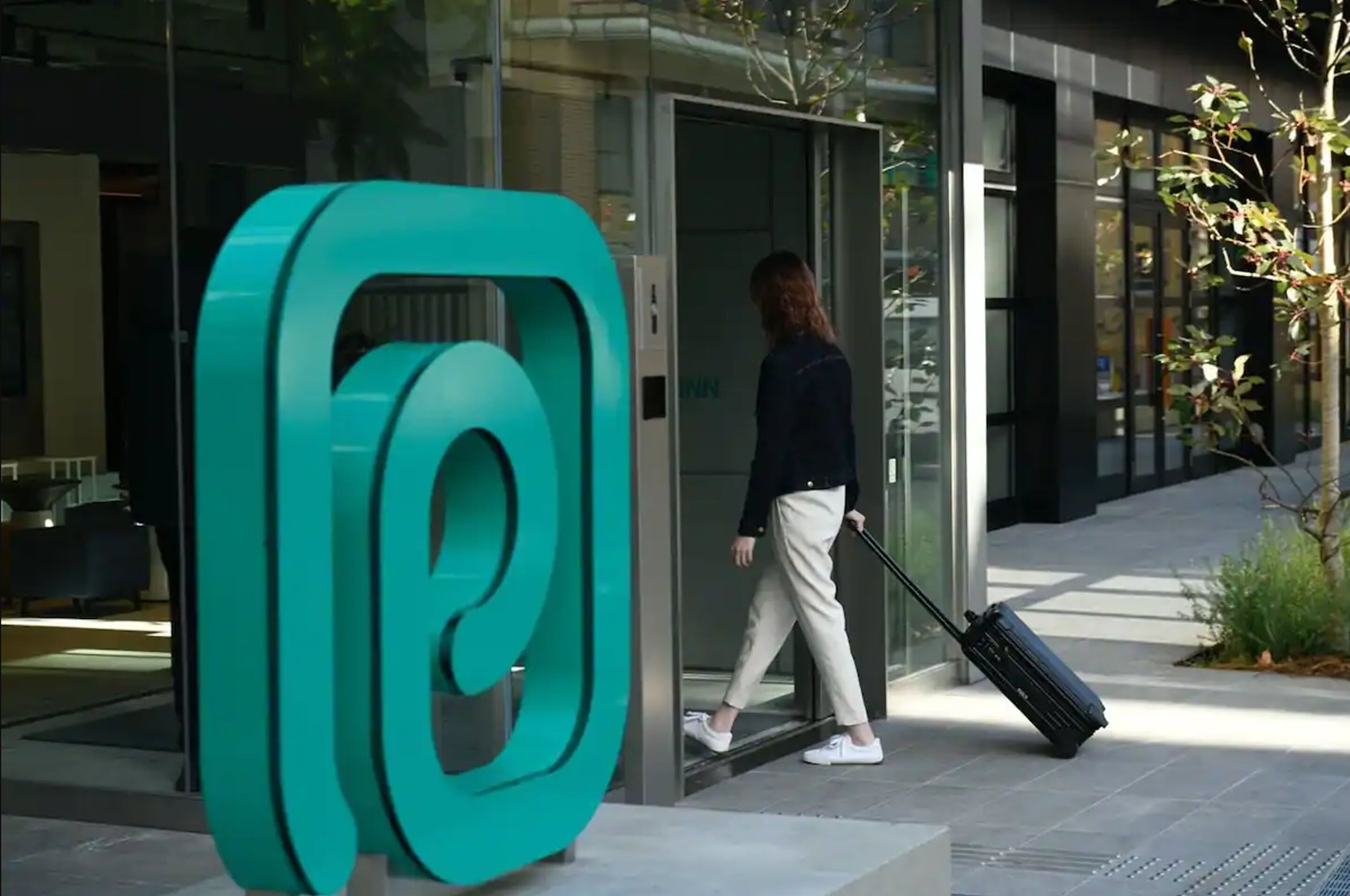 A modern building entrance with a large teal abstract sculpture, a glass door, and a person with a suitcase.