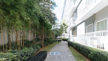 A pathway flanked by bamboo and shrubs, leading to a modern building with balconies.