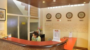 Zona de recepción de hotel con mostrador de recepción, un reloj que muestra la hora en San Francisco, Cebú, Tokio y París, y un menú colgado en la pared.