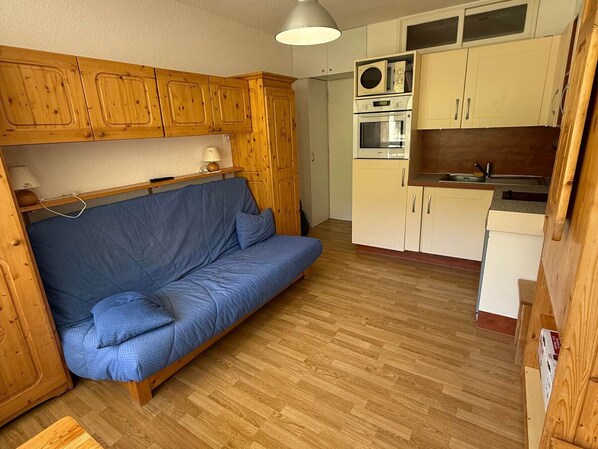 A compact living space with a sofa, wooden cabinets, a kitchenette, and a ceiling light fixture.