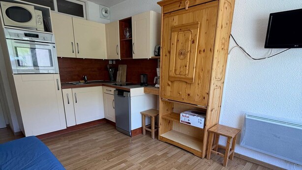 Una cocina compacta con armarios blancos, un microondas, un horno y un lavavajillas. Se observa un mueble de madera con un estante, un taburete pequeño y una caja etiquetada 'Tefal'. Un televisor está montado en la pared, y se ve un radiador.