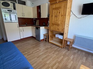 Pieni, käytännöllinen keittiö, jossa on valkoiset kaapit, mikroaaltouuni, uuni ja astianpesukone. Keittiössä on myös puinen kaappi, jossa on yksi hylly, pieni jakkara ja laatikko, jossa on merkintä ”Tefal”. Seinälle on asennettu televisio, ja lämpöpatteri on näkyvissä.
