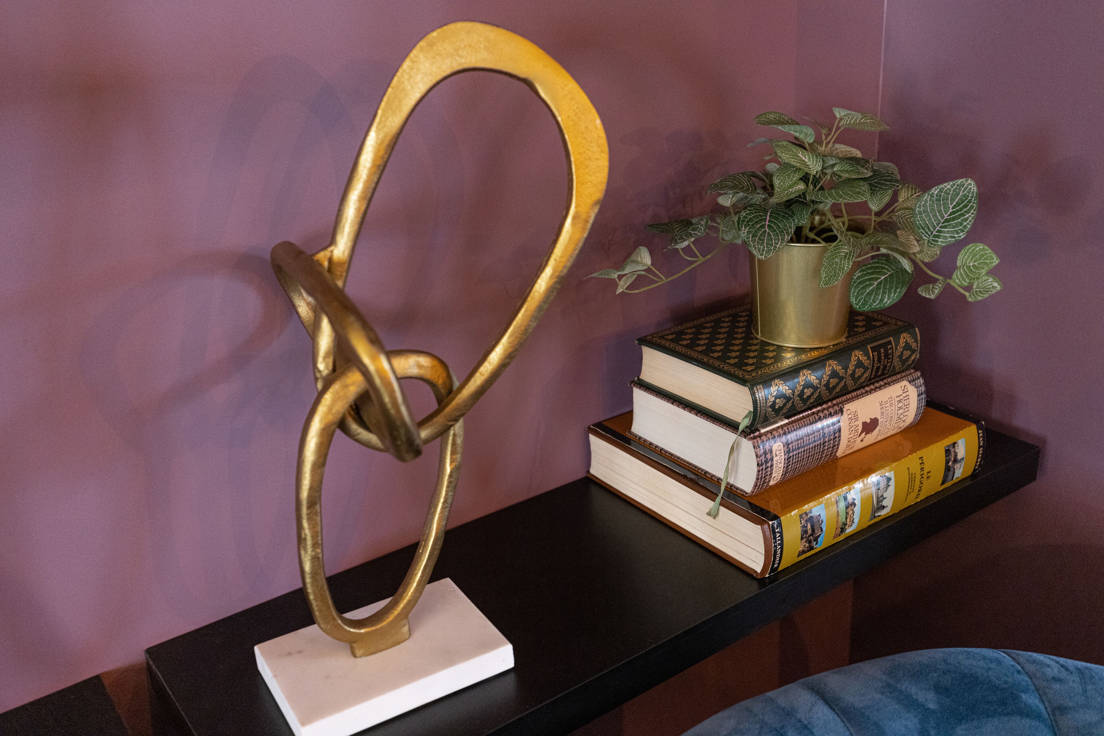 Una escultura metálica decorativa sobre un pedestal blanco, una planta en maceta y una pila de libros en un estante negro frente a una pared morada.