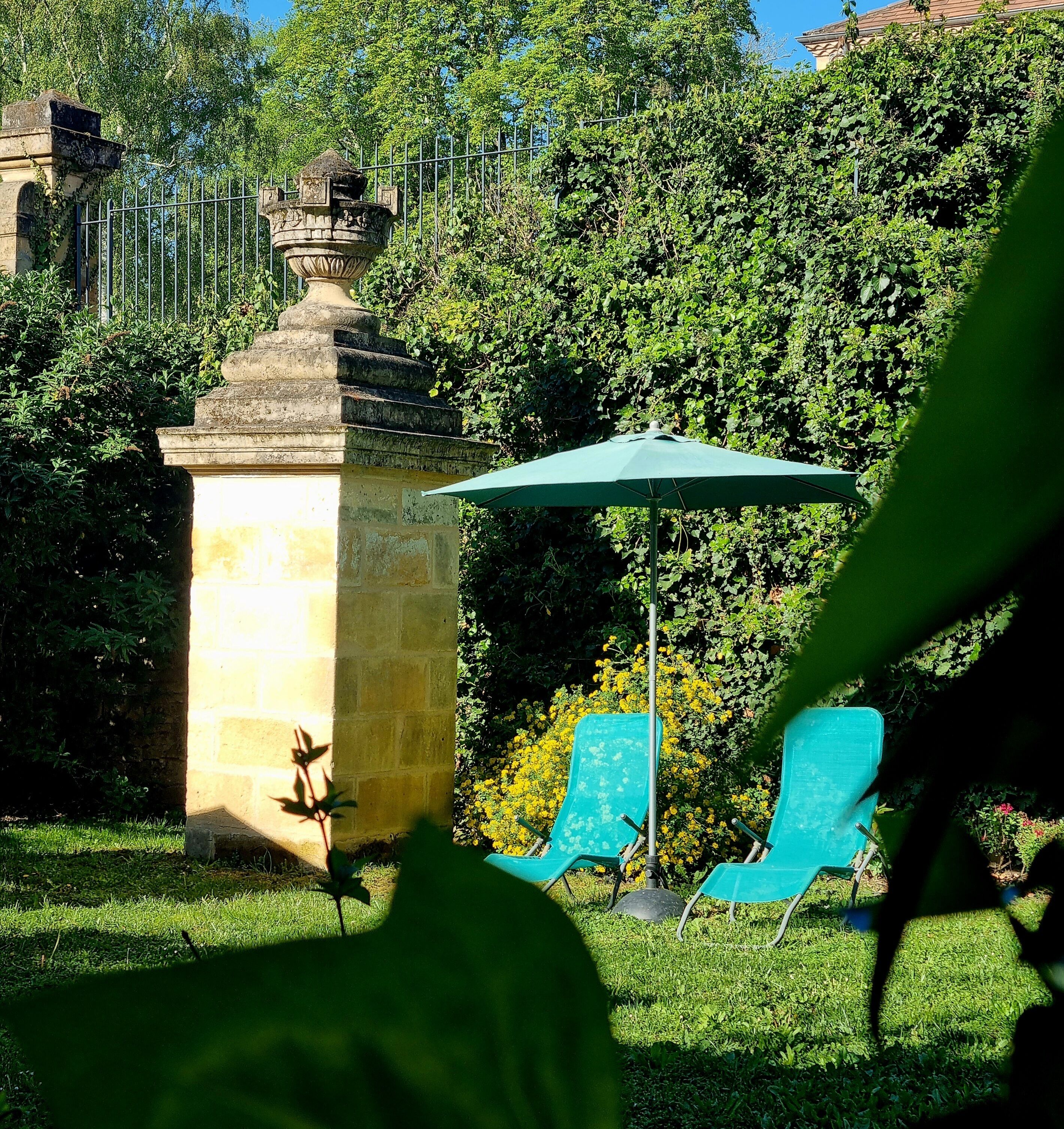 Un jardin agrémenté d’une structure en pierre, d’un parasol vert et de deux chaises bleues.