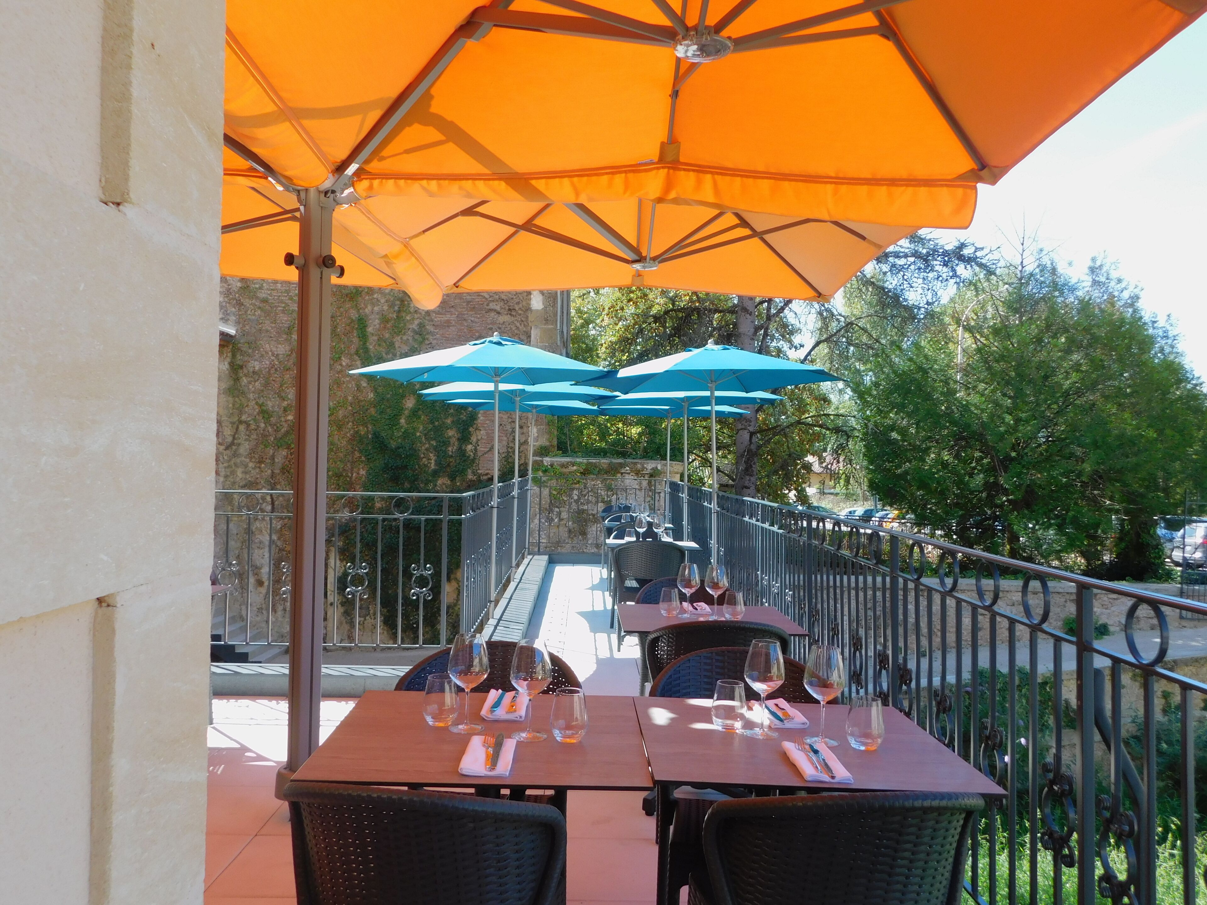 Un área de comedor al aire libre con mesas y sillas, cubierta por sombrillas naranjas y azules, y una barandilla de metal.