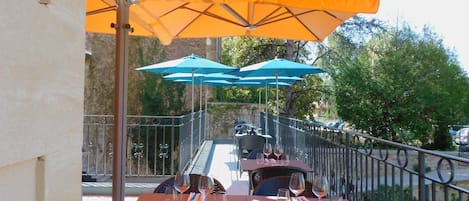 Un área de comedor al aire libre con mesas y sillas, cubierta por sombrillas naranjas y azules, y una barandilla de metal.