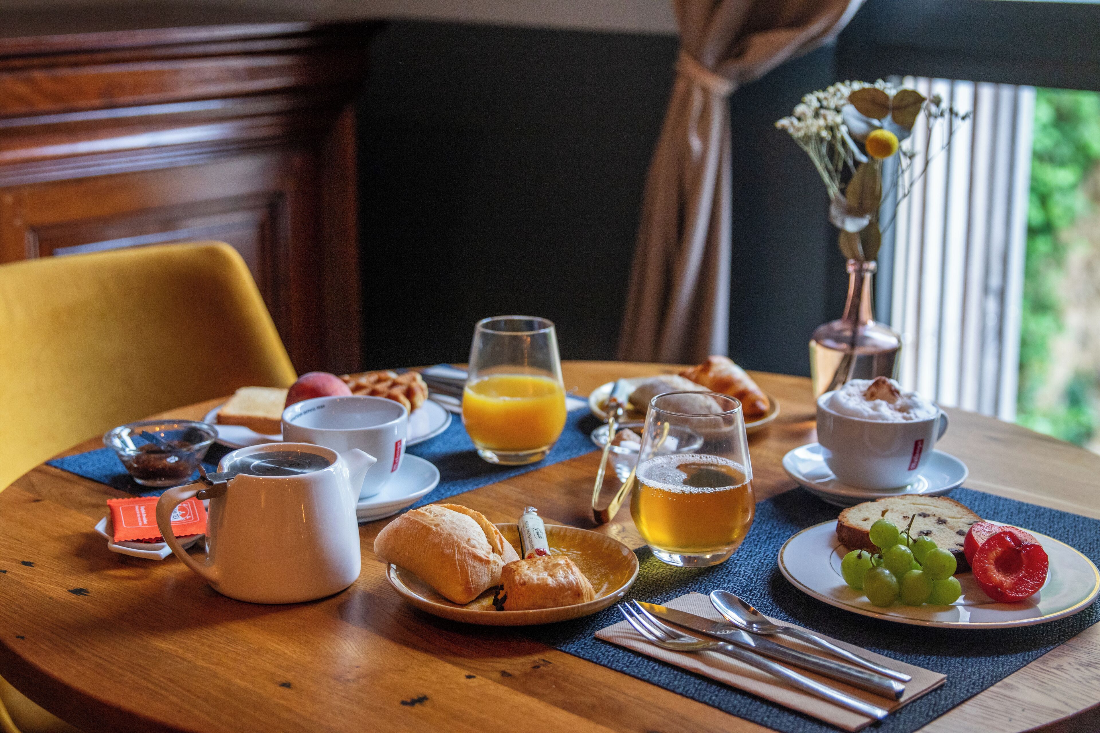 A breakfast table with croissants, a fruit plate, a coffee pot, a glass of orange juice, and a vase with flowers.