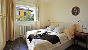 A bedroom with a bed, a nightstand, a lamp, a window with curtains, a radiator, and a wall-mounted picture. - Gravedona Aquamarine Apt. 3 (Gravedona)