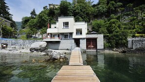 A modern house with a wooden dock leading to a stone entrance, surrounded by lush greenery and a clear water body. - Villa Parisi (Tremezzina)