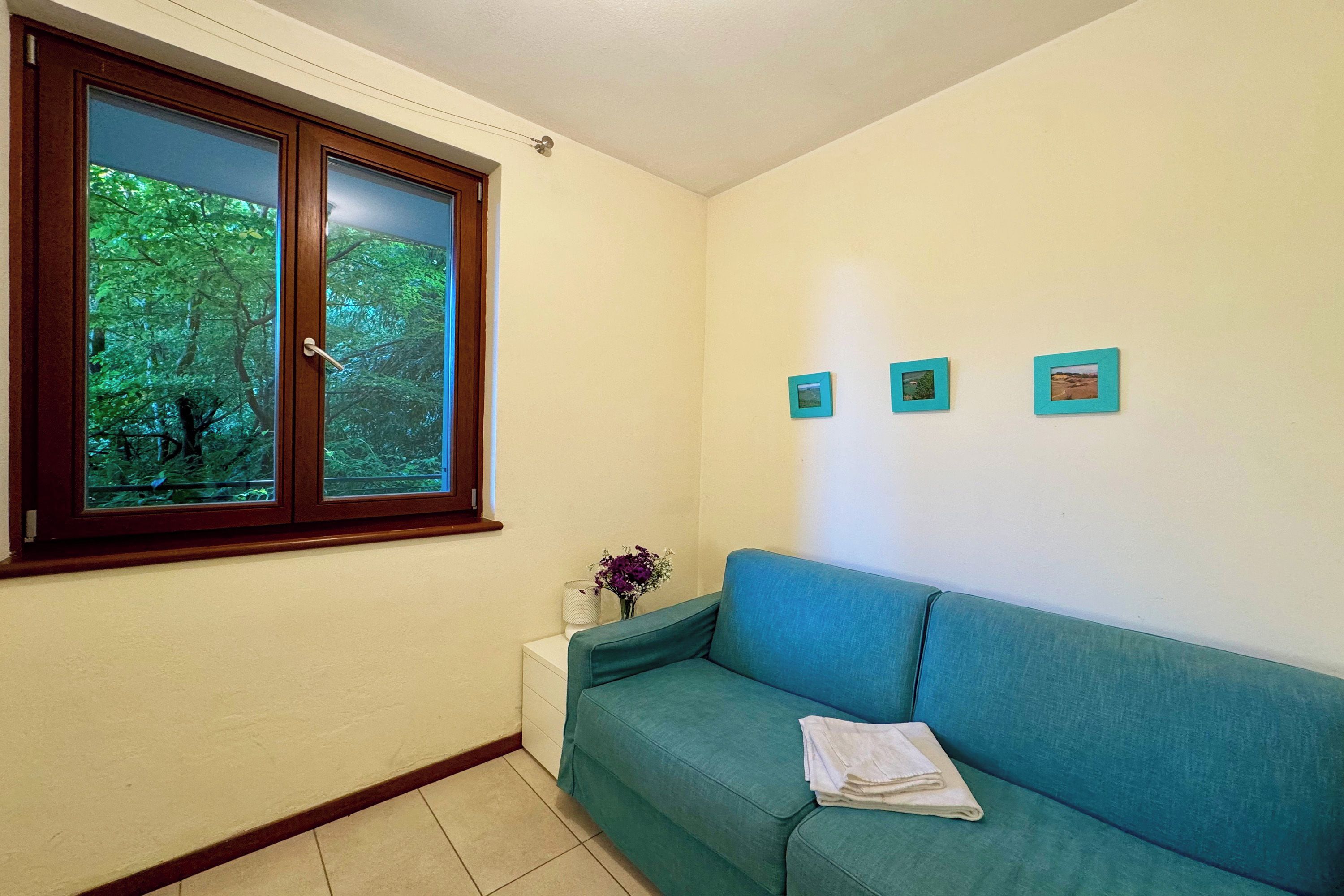 Ein Zimmer mit einem türkisfarbenen Sofa, einem Fenster mit Blick auf Bäume und drei kleine gerahmte Bilder an der Wand.