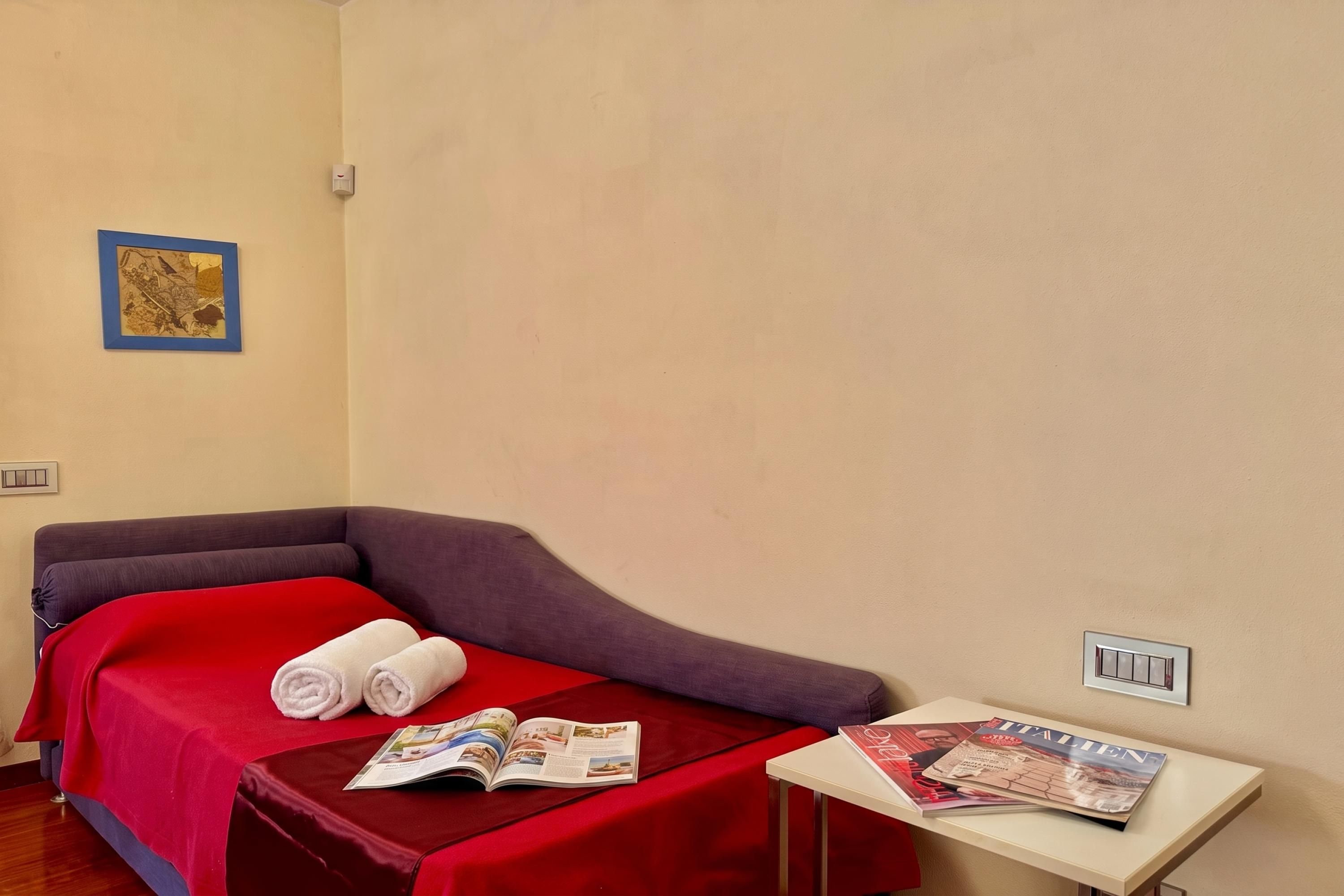 Un lit simple avec une couverture rouge, deux serviettes enroulées et une petite table avec des magazines.