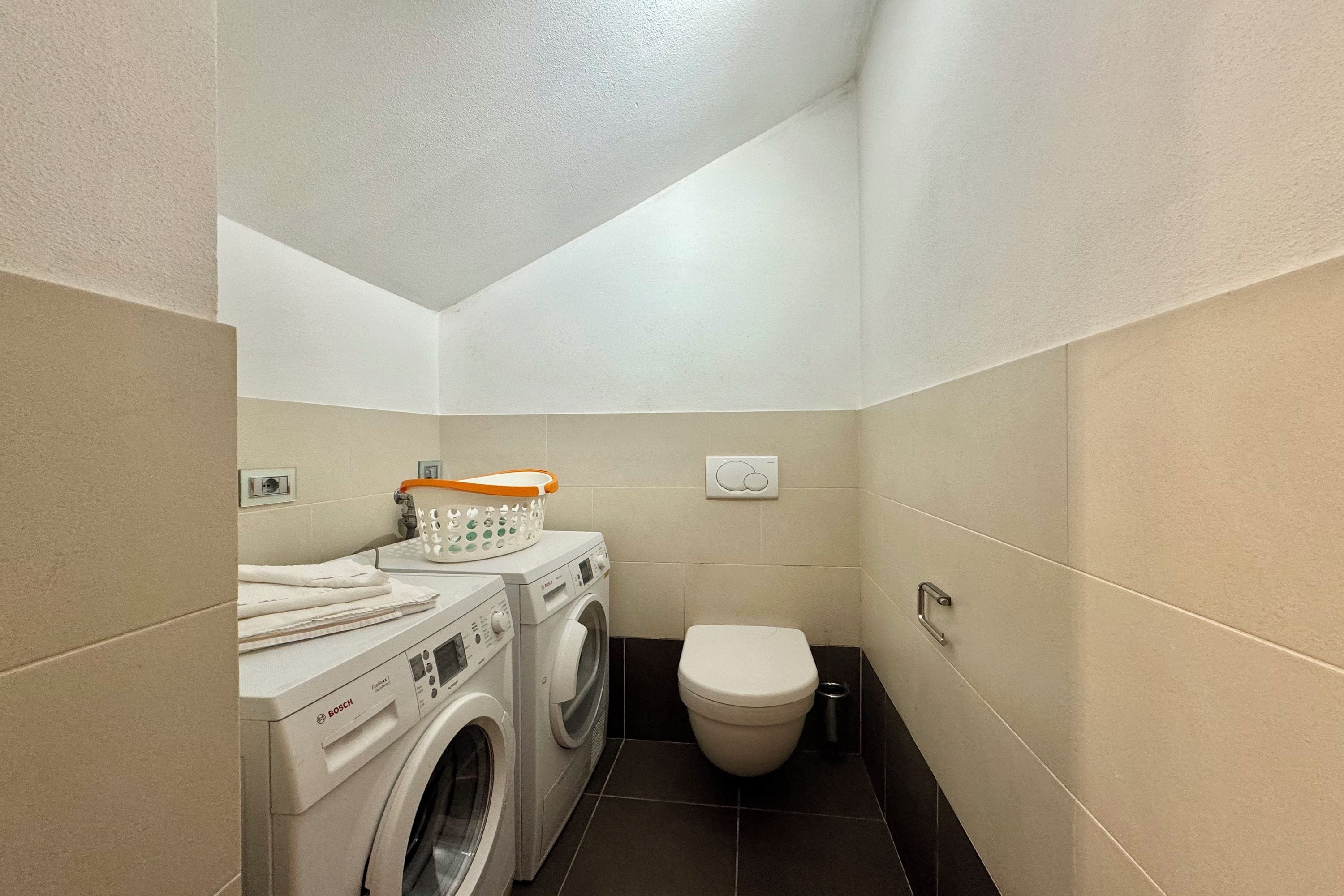 A compact laundry room with a Bosch washing machine and dryer, a toilet, and a small trash bin.