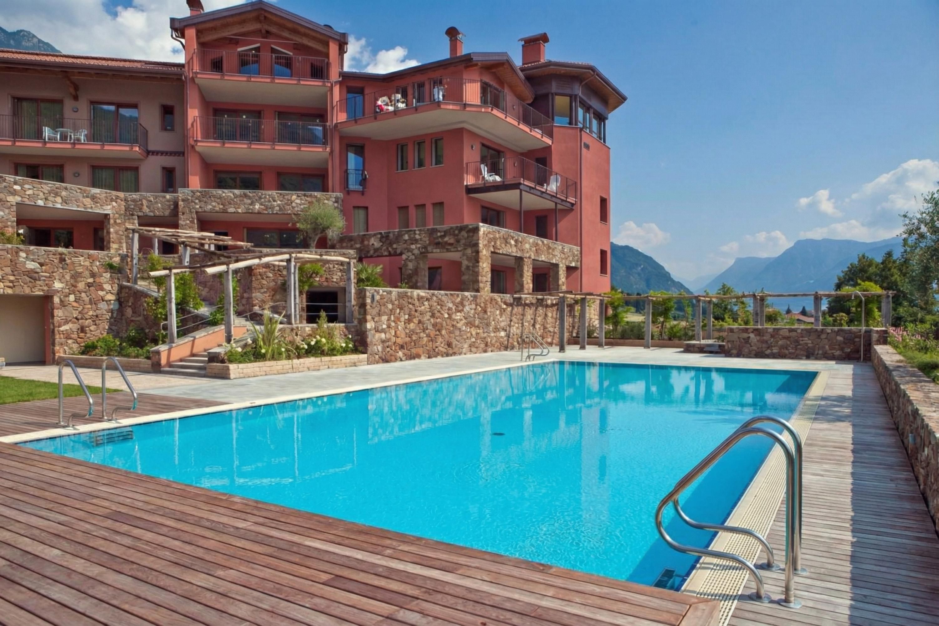 A pool area with a wooden deck, a stone wall, and a building with balconies.