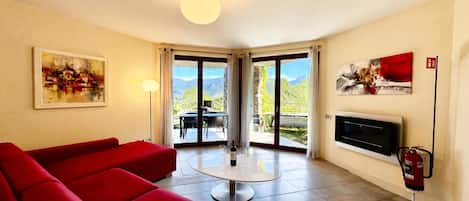 A modern living room with a red sofa, a round coffee table, and a fireplace. There are two large sliding glass doors leading to an outdoor patio with a table and chairs.