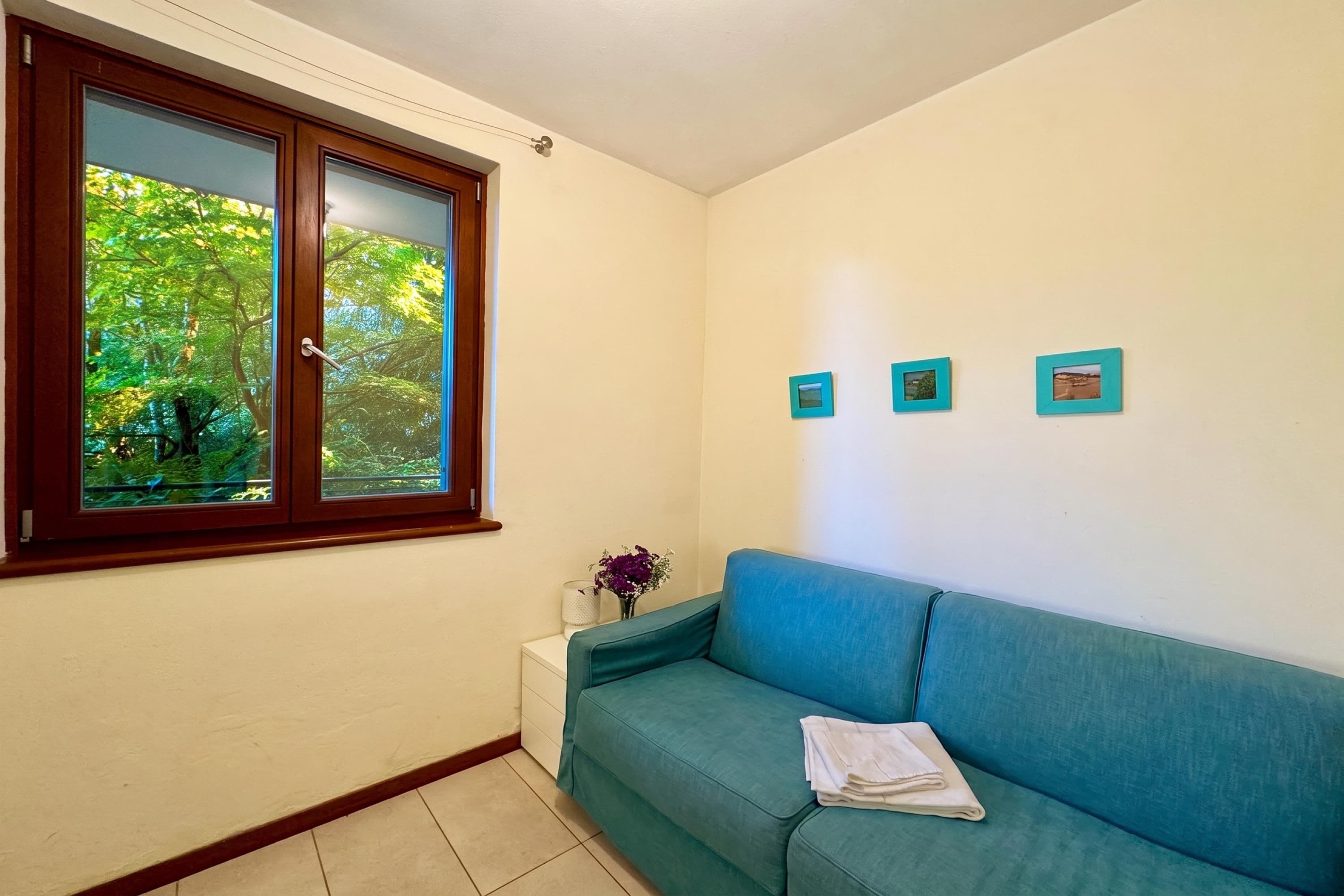 A room with a teal sofa, a small white side table with a vase, and three framed pictures on the wall.