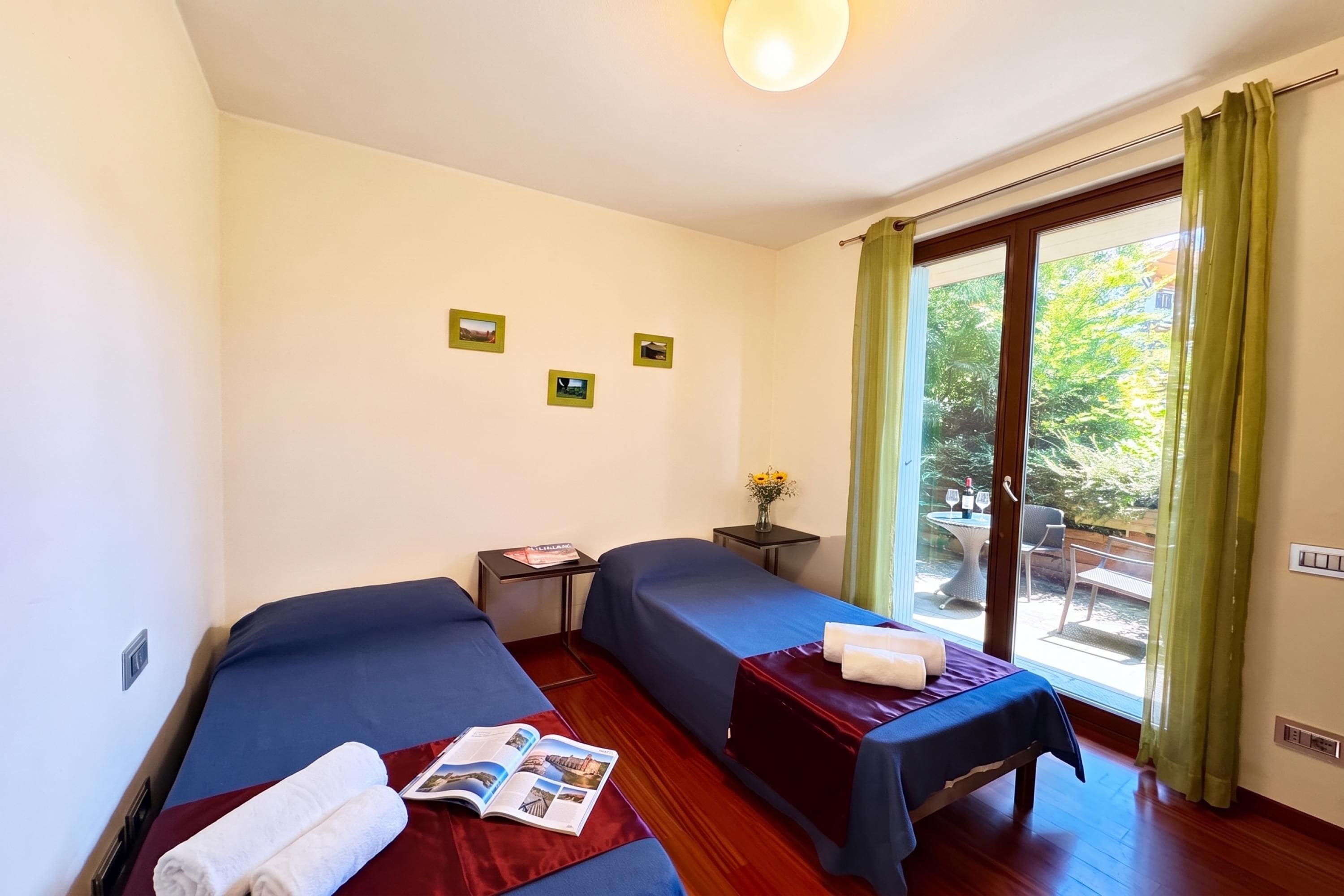 A hotel room with two beds, a small table with a magazine, a window with curtains, and a view of the outdoors.