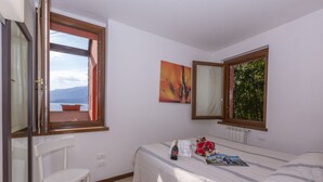 A bedroom with a bed, a chair, a window with a view, and a painting on the wall. - Fontana Del Lago Apt. 8 (Varenna)