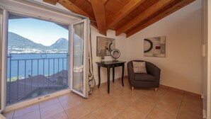 A room with a balcony overlooking a lake and mountains, a small table with a lamp, and a sofa. - Casa Mastena Vista Lago (San Siro)