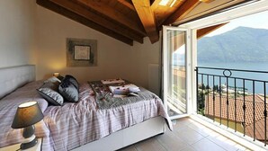 A bedroom with a bed, a bedside lamp, a window with a view of mountains, and a balcony with a railing. - Casa Mastena Vista Lago (San Siro)