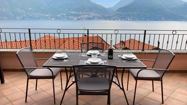 A dining table set for four with chairs, a view of a lake and mountains, and a balcony railing. - Casa Mastena Vista Lago (San Siro)
