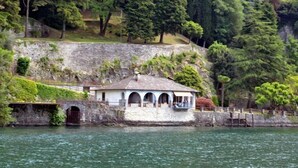 A small, white building with a tiled roof and arched windows, situated by a body of water with lush greenery in the background. - Villa Chez Nous (Pognana Lario)