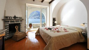 A bedroom with a stone fireplace, a bed with rose petals, and a view of the sea. - Villa Chez Nous (Pognana Lario)
