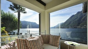 A room with a view of a lake and mountains, a cushioned bench with striped pillows, and a small table with a book. - Villa Chez Nous (Pognana Lario)