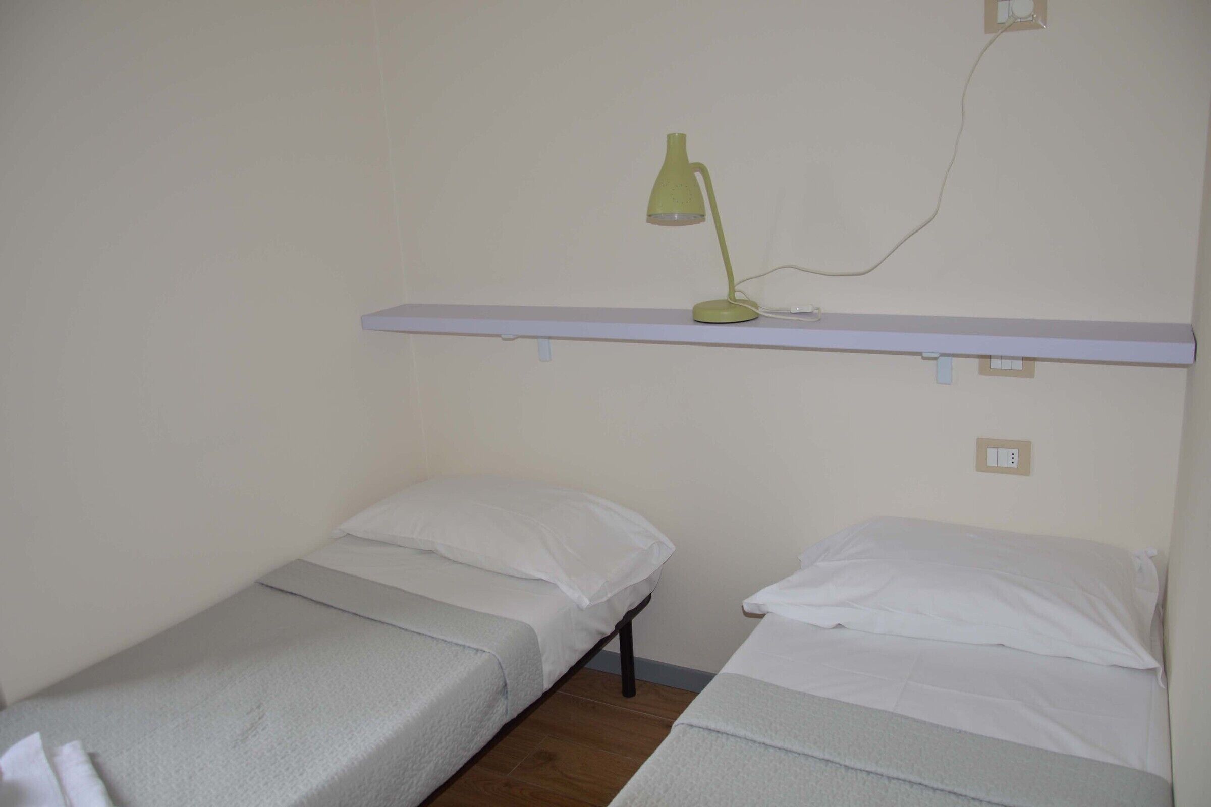 A small, simple room with two single beds, a white shelf above, and a green desk lamp.