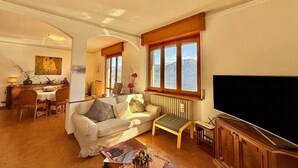 A living room with a sofa, a coffee table, a TV, and a view of mountains through large windows. - Isola Vista - Terrazzo (Sala Comacina)