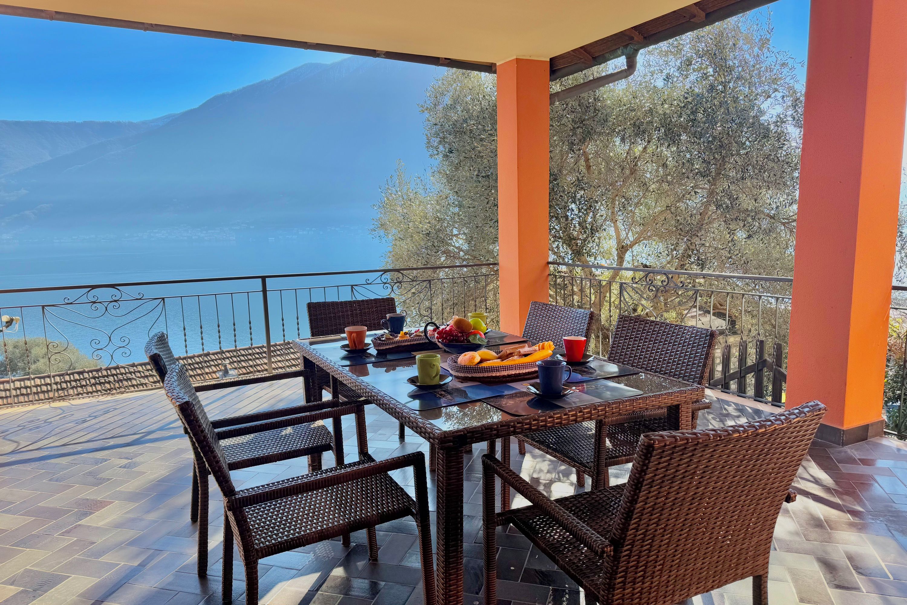 A terrace with wicker furniture, a table set with fruits and cups, and a view of a lake and mountains.